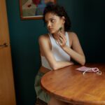 A young woman using a jade face roller sitting at a wooden table indoors.